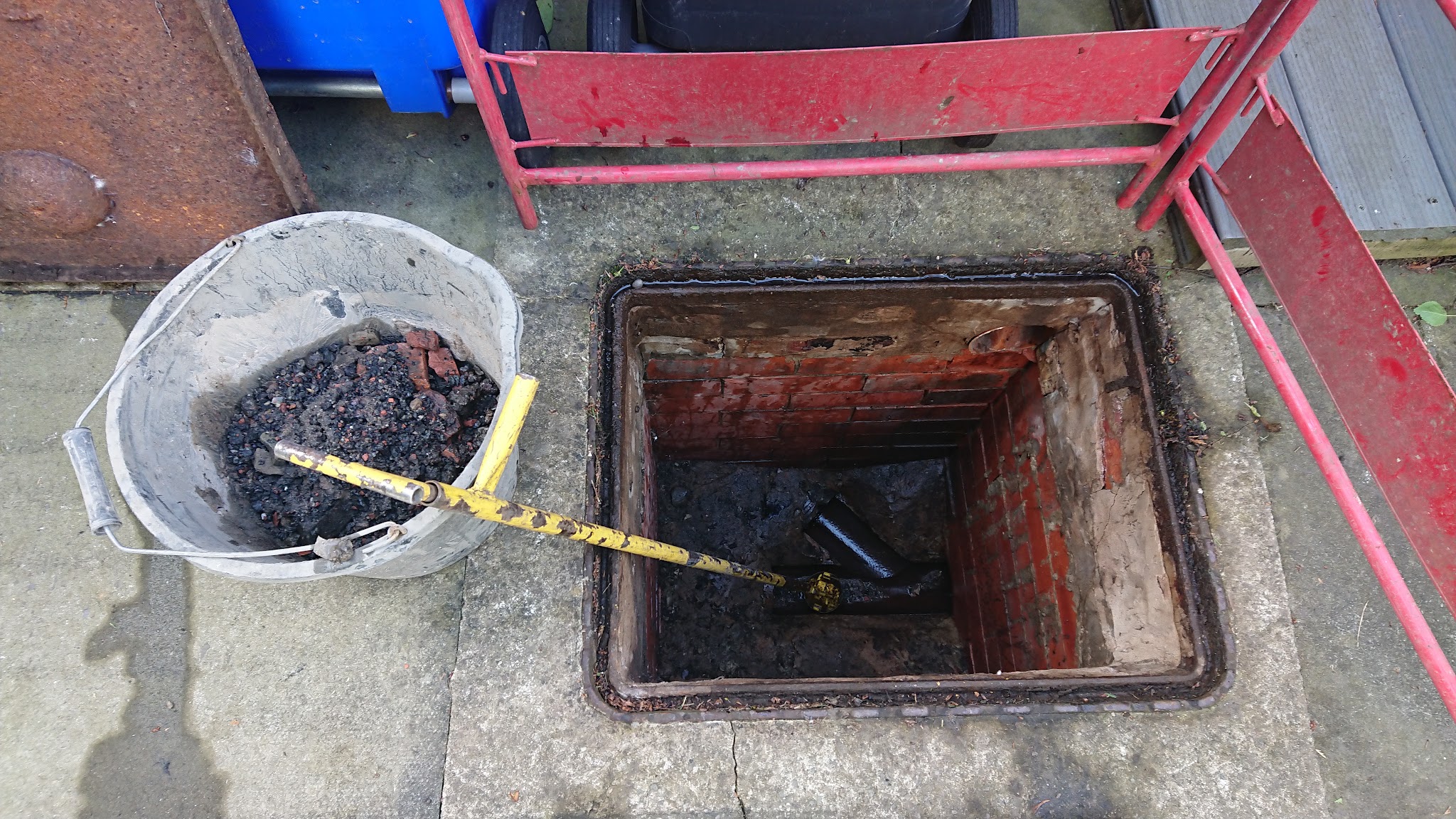 An opened brick drain chamber with a rod and a bucket of debris beside it