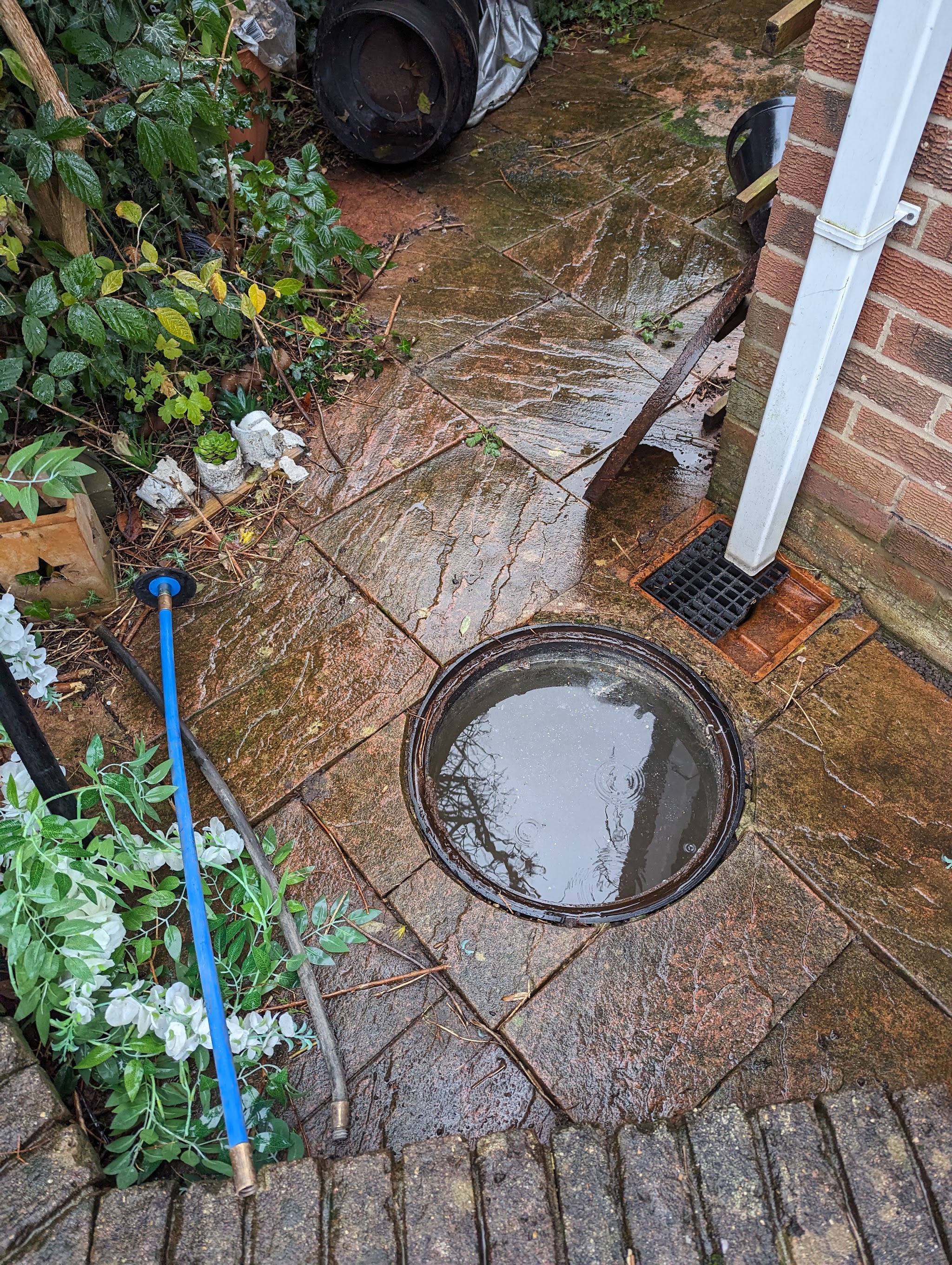 An open drain chamber on a paved patio with a drain rod laid beside it