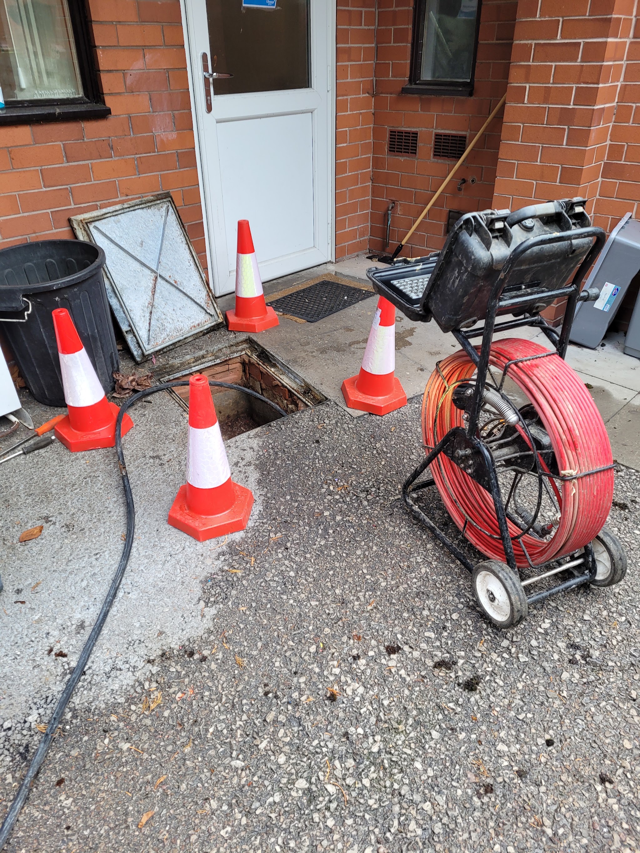 A CCTV drain survey reel and monitor beside an open chamber, with traffic cones around it