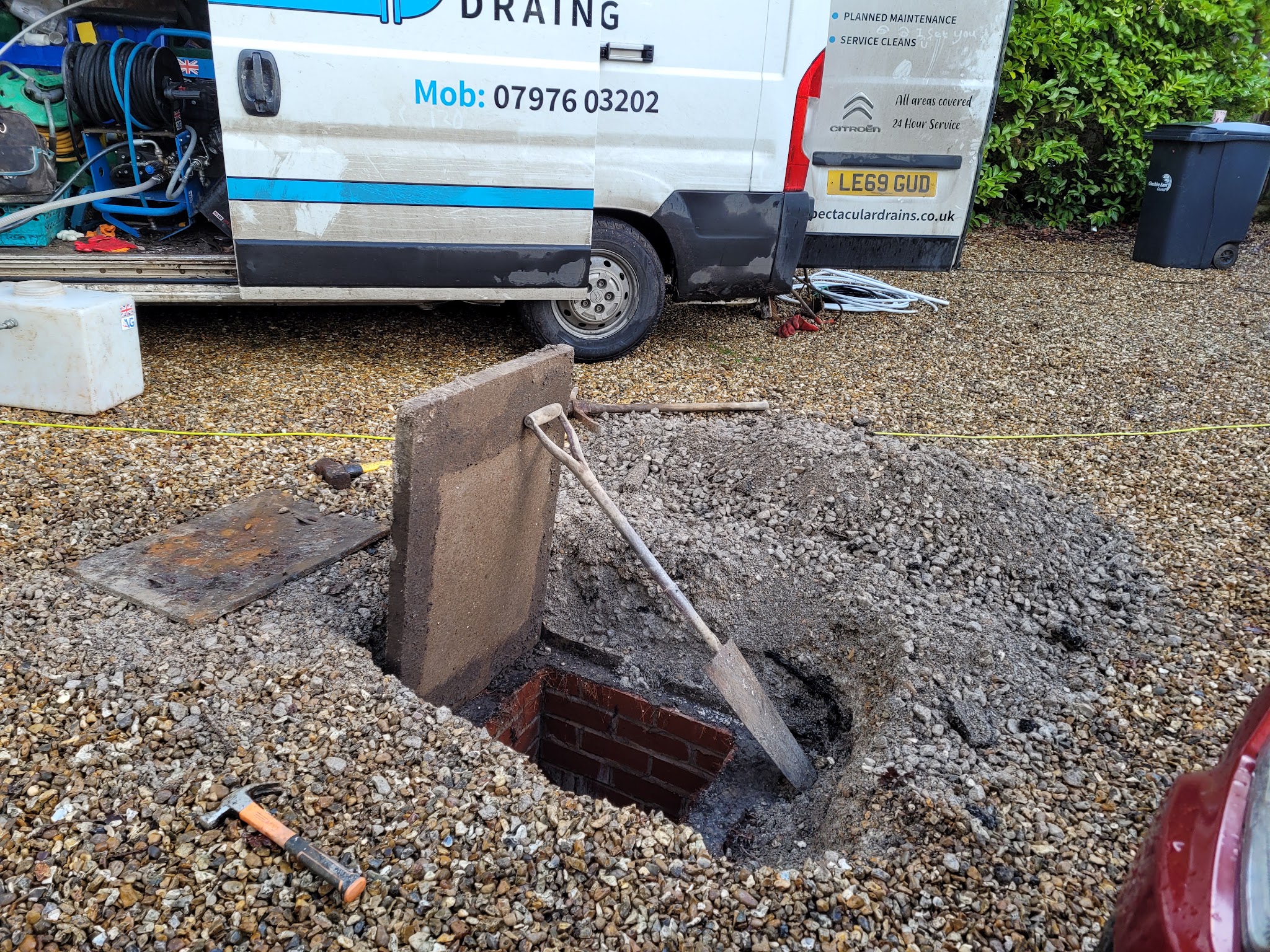 The van with its back doors open beside an excavated drain chamber on a gravel drive